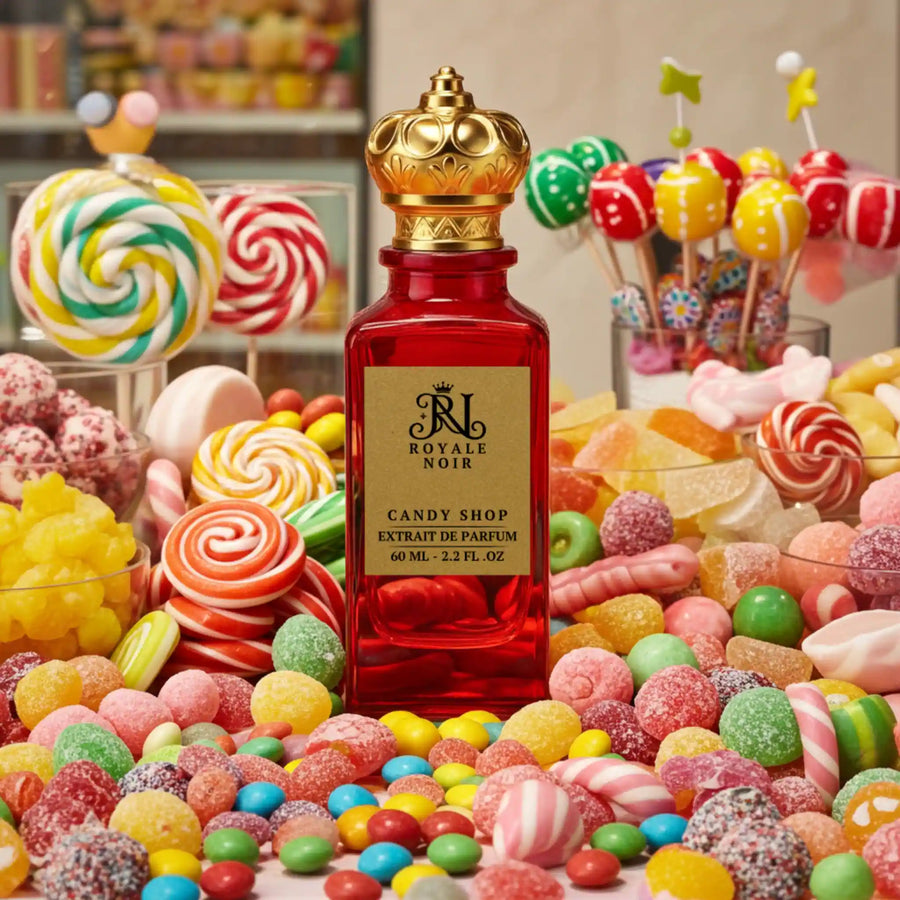 A luxurious red glass perfume bottle with a gold crown-shaped cap, labeled “Royale Noir – Candy Shop Extrait de Parfum, 60 mL – 2.2 FL. OZ.” The bottle is arranged on a rich burgundy background surrounded by candy and gourmand ingredients: assorted colorful jelly beans, marshmallows, pink chewing gum, caramel candies, pieces of brown sugar, vanilla beans, cacao beans, white jasmine flowers, wood sticks, a small bowl of white powder, dark dried fruits, green leaves, and a purple violet flower.