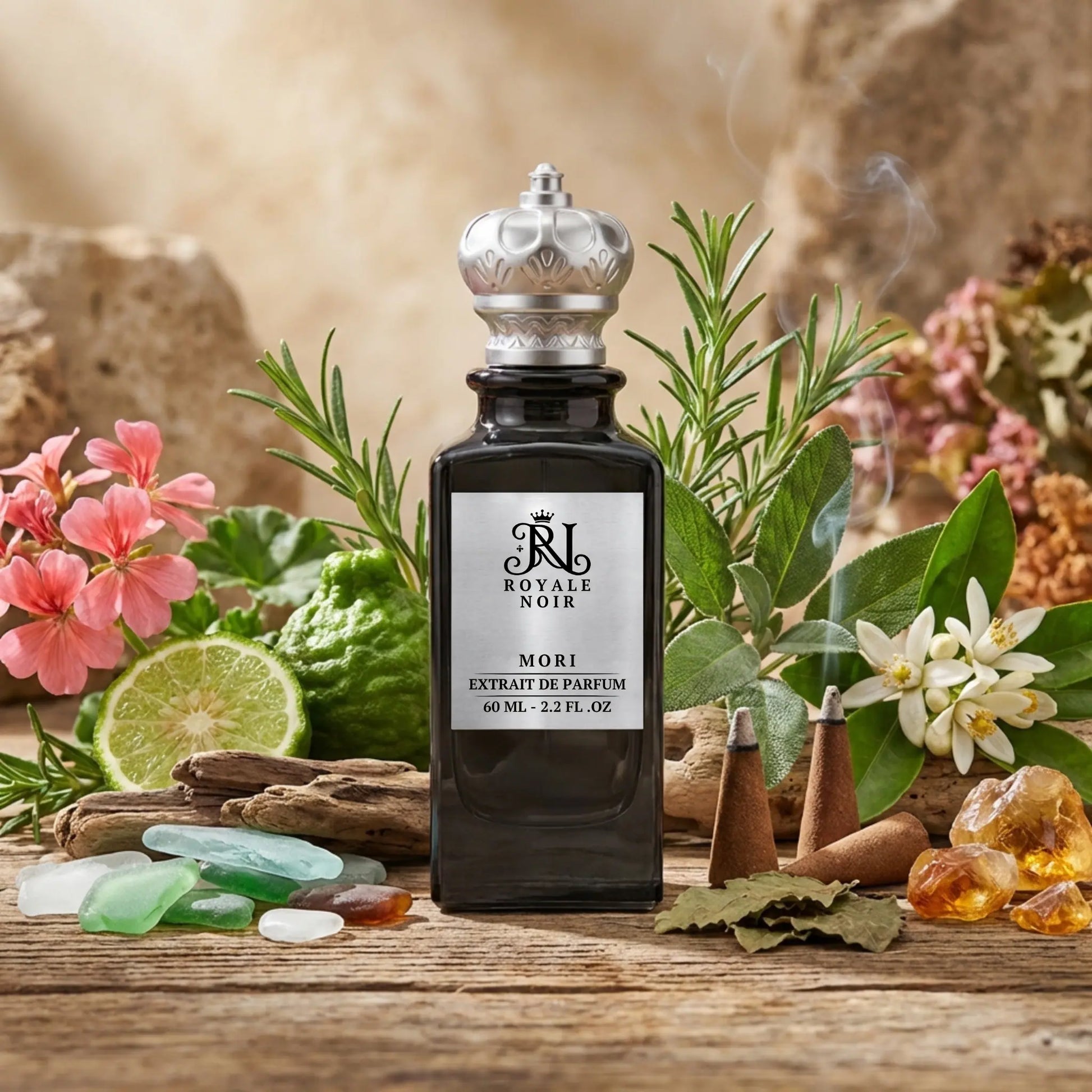 A black perfume bottle labeled "Royale Noir Mori Extrait de Parfum, 60 ml - 2.2 fl. oz" is centered on a wooden surface. The bottle has an ornate silver cap. Surrounding the bottle are fragrant ingredients, including a slice of lime, rosemary, pink and white flowers, mint leaves, sage leaves, coarse salt, and resin pieces. The composition evokes a sense of luxury and botanical richness.