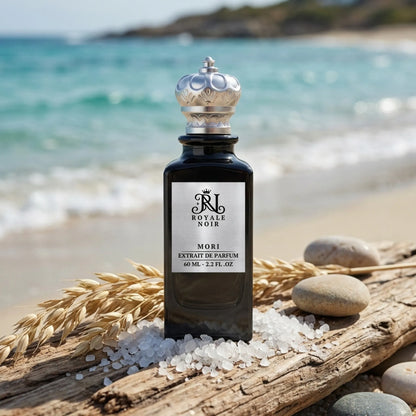 A black perfume bottle labeled "Royale Noir Mori Extrait de Parfum, 60 ml - 2.2 fl. oz" on a beach with ocean in the background